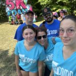 Group selfie of smiling volunteers in blue shirts outdoors, with a bright event banner and trees in the background.