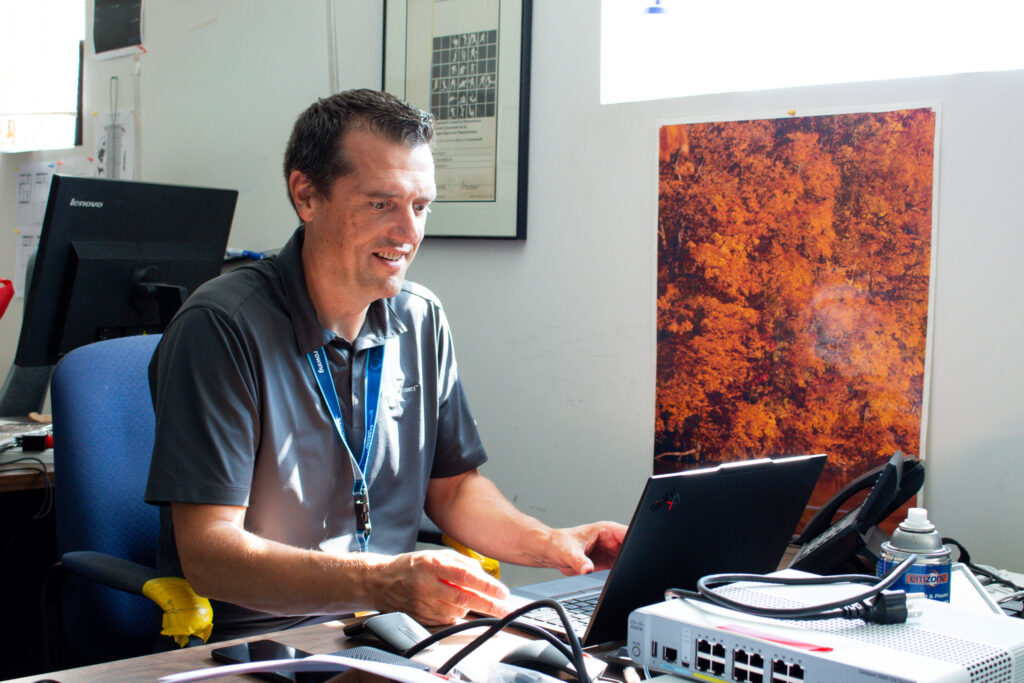 Man in dark polo working at a desk with two laptops and network equipment, smiling at the screen.