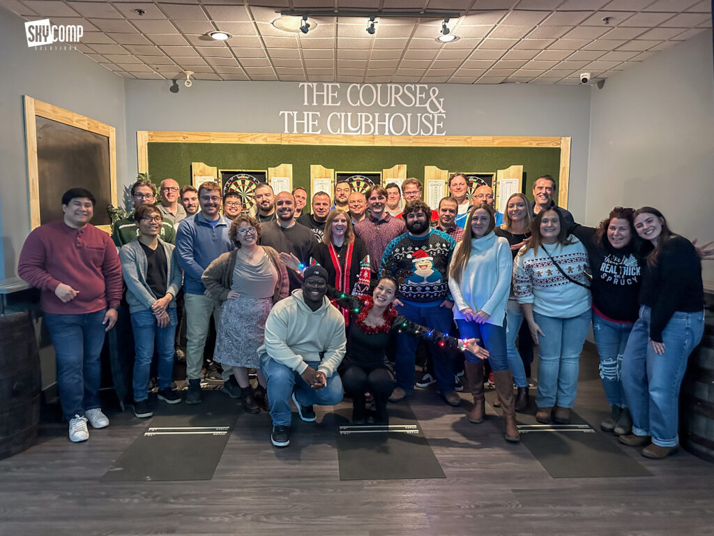 Group of coworkers posing for a team photo in a game room with dart boards and a 'THE COURSE & THE CLUBHOUSE' sign in the background.
