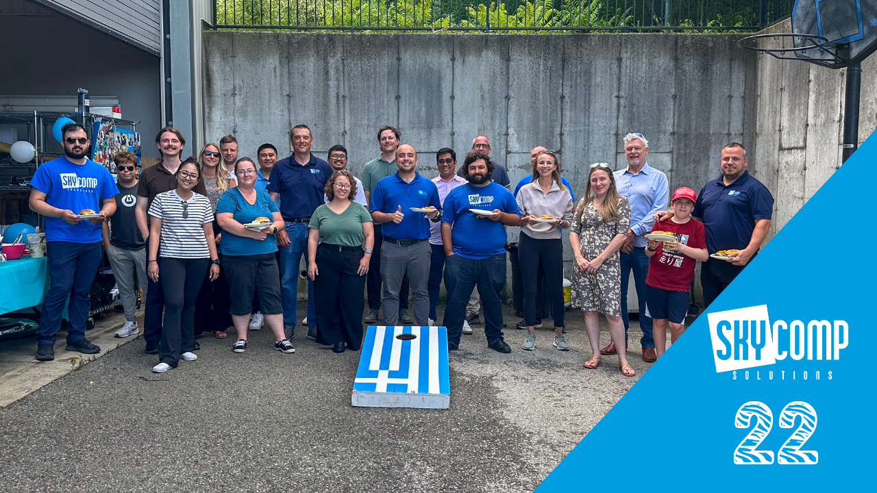 Group of SkyComp Solutions employees posing outdoors for a team photo at a company gathering, some holding plates of food, with a blue corner design on the right.