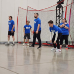 Five people in blue shirts stand near a red net barrier inside an indoor gym, watching an activity.