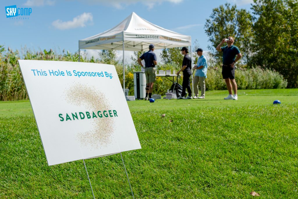 Sponsorship sign on a golf course reading 'This Hole Is Sponsored By: SANDBAGGER' with a white canopy and people in the background.