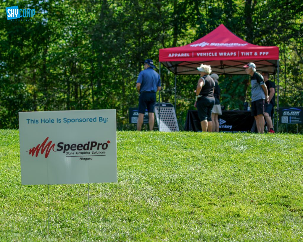 Outdoor golf event scene with a sponsor sign in the foreground reading 'This Hole Is Sponsored By: SpeedPro' and a red canopy in the background.