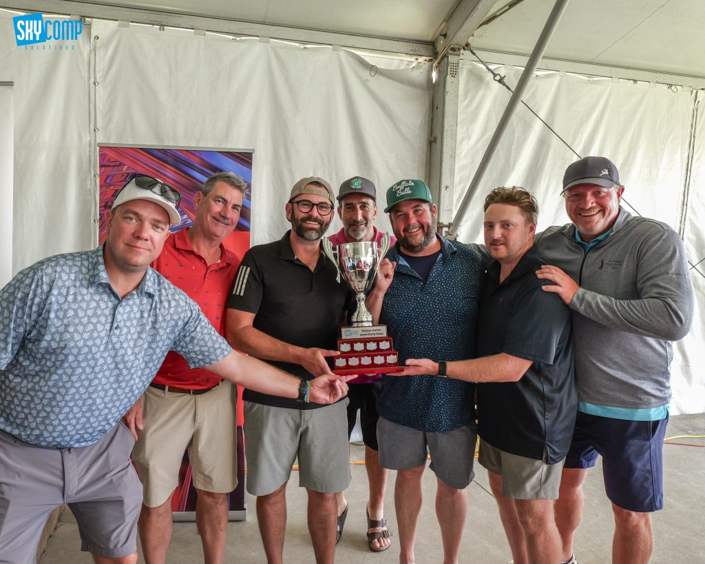 Six men in casual shirts stand in a white tent, posing with a large trophy for a charity event.