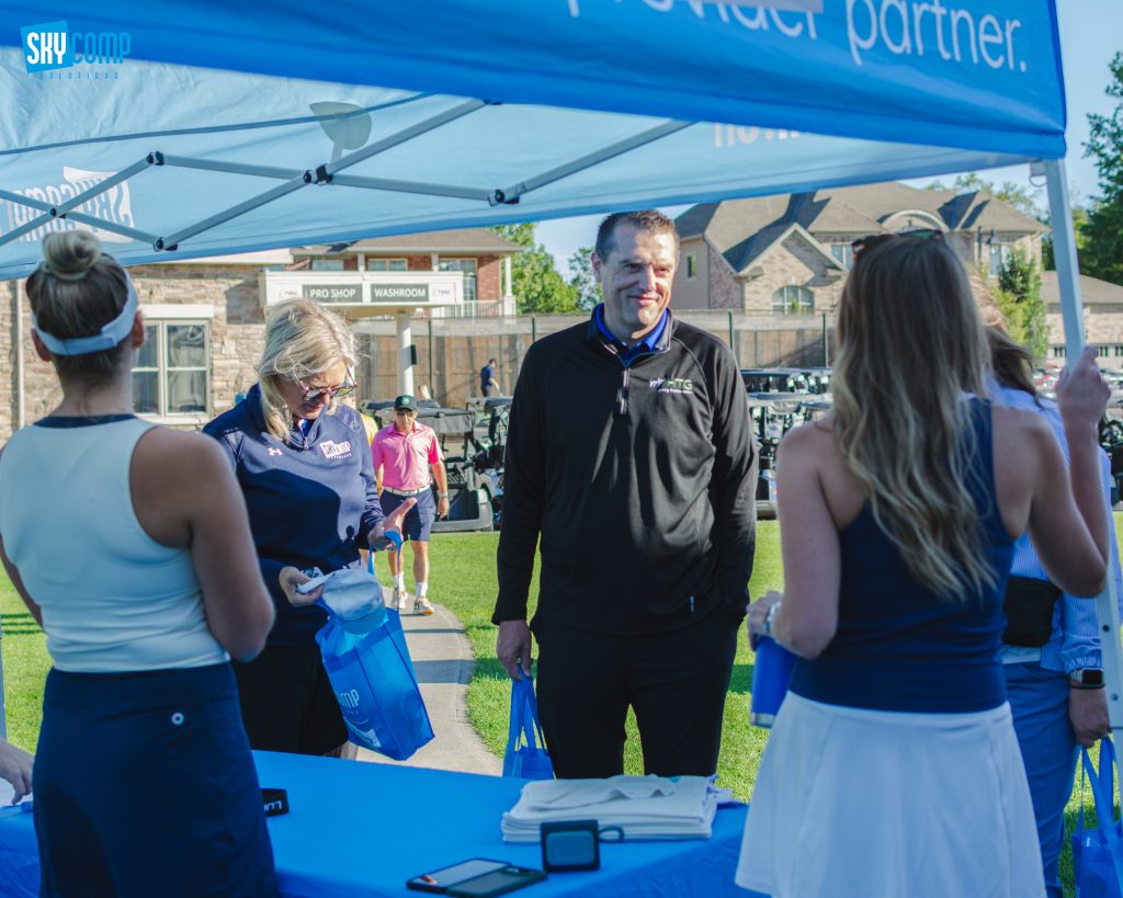 Volunteers under a blue canopy at an outdoor event, with a registration table and blue tote bags nearby.
