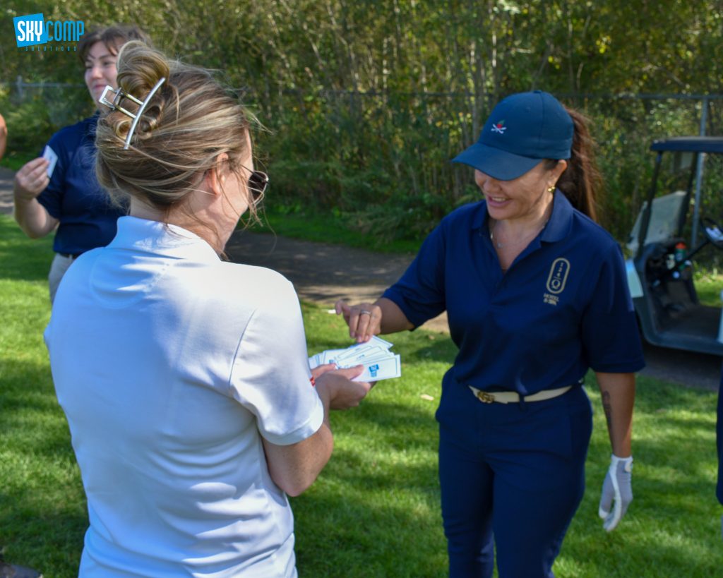 Two women outdoors exchanging playing cards, one in a blue uniform and the other in a light shirt; SkyComp logo in the top-left corner.
