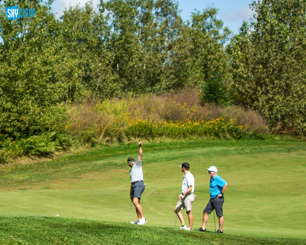 Group of golfers on a sunny green course with trees in the background; a player raises his arm in celebration near the foreground; SkyComp logo in the top-left corner.